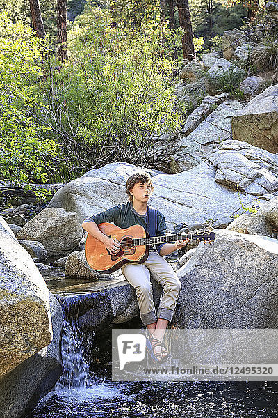 Ein junger Mann spielt nachdenklich auf seiner Gitarre  während er auf den Felsen entlang eines Baches sitzt/steht.