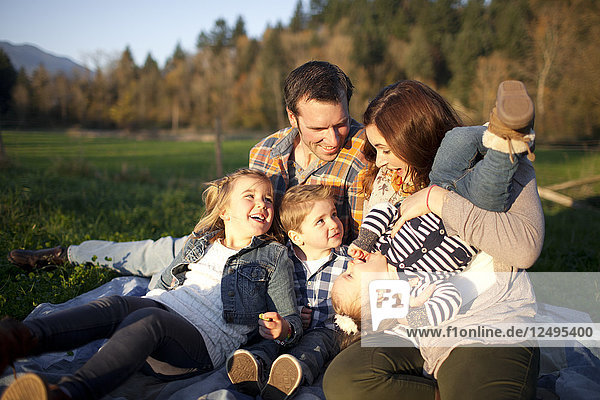 A family of five laugh together while sitting on a blanket outdoors.