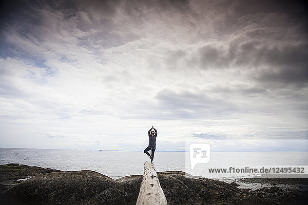 A young woman does the Yoga Tree Pose while balancing on a log at the beach near Sechelt  BC  Canada.