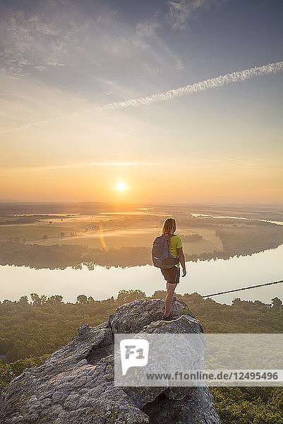 Frau steht auf einem Sandsteinüberhang und betrachtet den Sonnenaufgang vom Gipfel des Petit-Jean Mountain über dem Arkansas River Valley