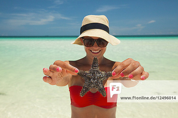 A young woman wearing a bikini and sun hat holds up a starfish at the beach in Cuba.