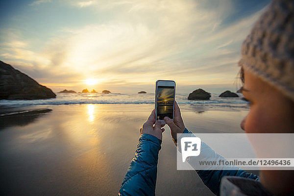 Eine junge Frau macht mit ihrem Smartphone ein Foto vom Sonnenuntergang am Strand von Bandon  Oregon.