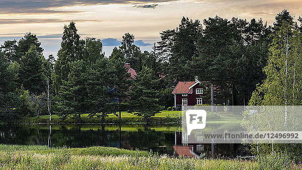 typisch schwedisches Haus  das sich im Wasser eines Sees in der Nähe von Mora in Schweden im Sommer spiegelt  mit Bäumen rundherum und einem schönen Himmel mit Wolken und schönem Sonnenuntergangslicht.