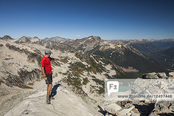 A hiker points down the valley at an alpine lake from the summit of Cassiope Peak near Pemberton  British Columbia  Canada.