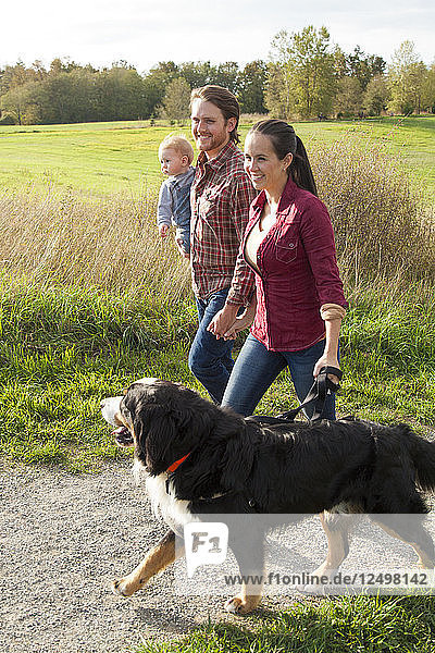 Ein junges  attraktives Paar geht mit seinem Baby und seinem Berner Sennenhund auf einem Kiesweg im Park spazieren.