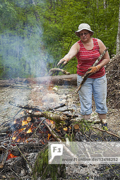 Eine Frau mittleren Alters wirft bei der Gartenarbeit einen Ast auf ein großes Feuer.