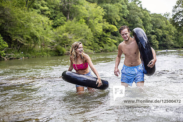 A young man and woman talk and laugh while walking with tubes down a river.