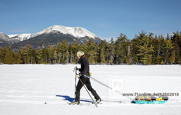 A Man Cross Country skies across a frozen lake while pulling a pulk sled. In the background can be seen Mount Katahdin.
