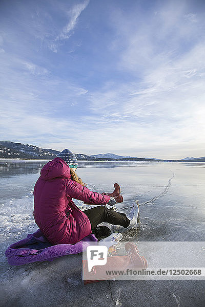 Ein Teenager-Mädchen  das auf dem gefrorenen Pend Oreille River sitzt und seine Schlittschuhe anzieht