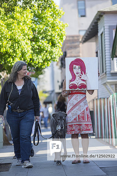 A woman holding a bust-up painting in front of herself in San Diego's Little Italy as a passersby and her dog curiously look on.