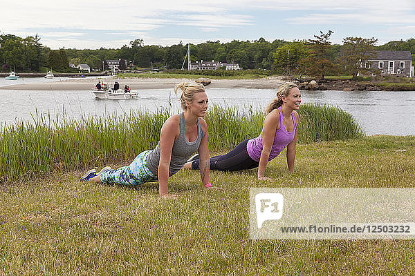 Two women in upward dog pose doing yoga outside on the grass near the water