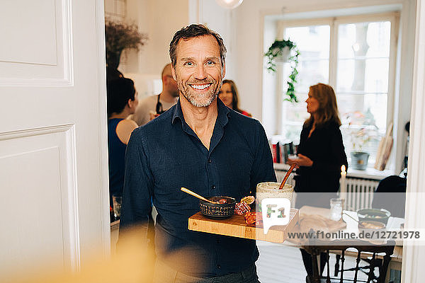 Portrait of smiling mature man holding food in serving tray while standing against friends at party