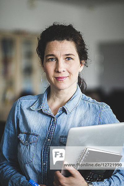 Portrait of confident mature female teacher holding diary and laptop while standing at computer lab