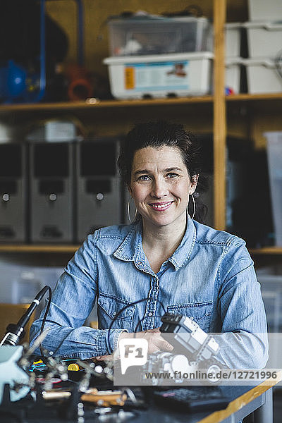 Portrait of confident mature female teacher sitting with science project at desk in classroom at high school