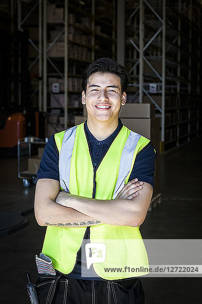 Portrait of smiling young male warehouse worker standing with arms crossed at storage room