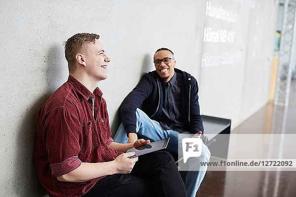 Smiling student holding digital tablet while sitting with friend on bench in university