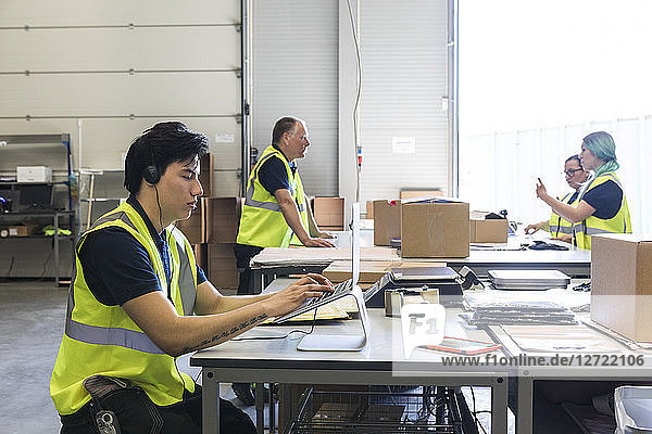 Confident young male customer service representative using laptop while coworkers discussing at desk in warehouse