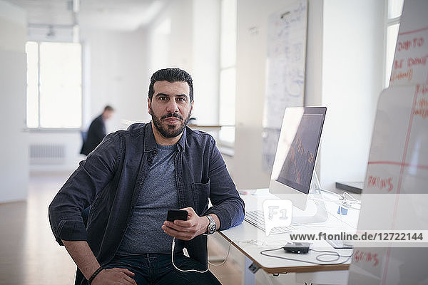 Portrait of male engineer holding mobile phone while sitting at computer desk in office