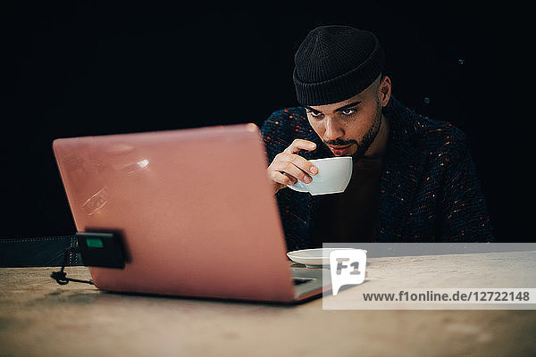 Confident young male hacker drinking coffee while looking at laptop on desk in office