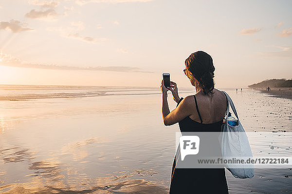 Rear view of woman photographing sunset over sea using mobile phone at beach