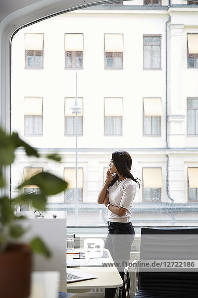 Side view of mature woman talking on mobile phone while looking through window at office