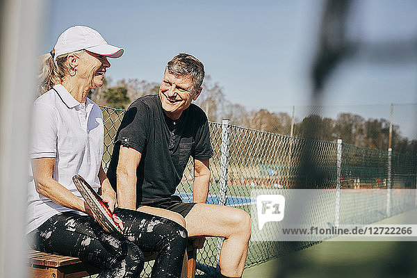 Happy senior man and woman talking while sitting on bench at tennis court