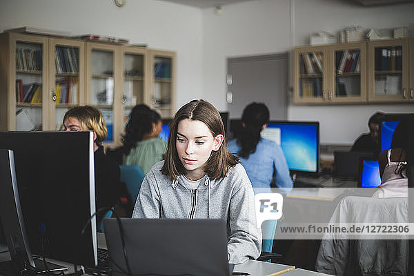 Confident female student using laptop at desk against teacher and friends sitting in computer lab