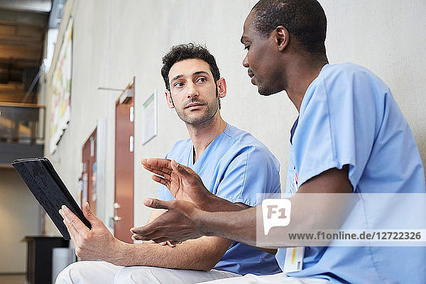Male nurse gesturing while discussing over digital tablet with coworker in corridor at hospital