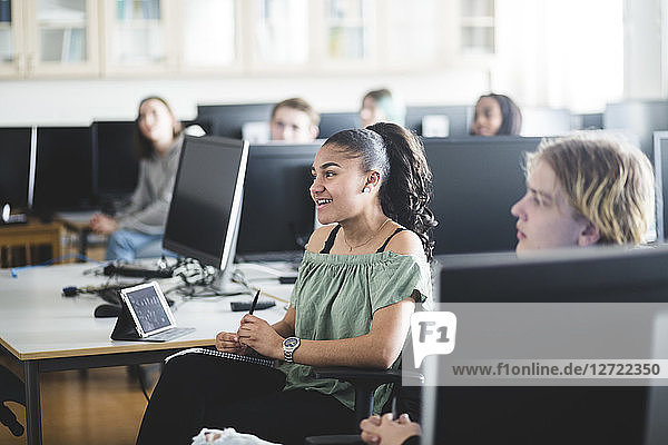 Smiling teenage girl sitting by young friend amidst computers at lab in high school
