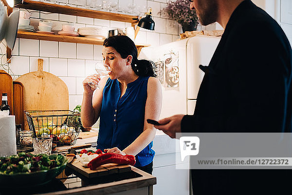 Male friend looking at mature woman drinking wine while standing in kitchen