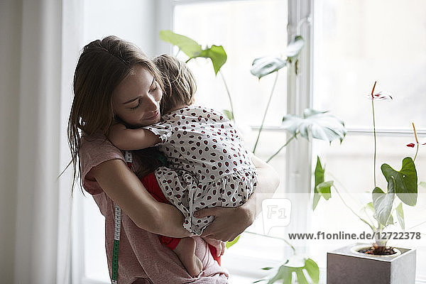 Female fashion designer embracing daughter while standing by window at home