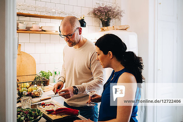 Woman with wineglass looking at male friend cutting pepper in kitchen