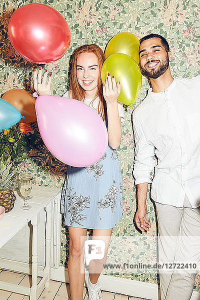 Portrait of smiling young woman playing with balloons while standing by man against wallpaper at home during dinner part