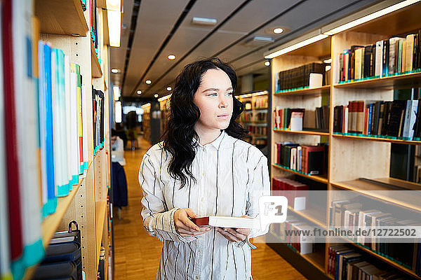 Young female student holding book while standing in university library
