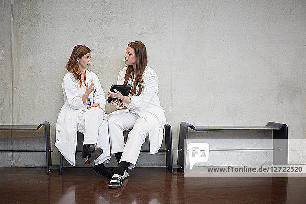 Full length of confident female doctors discussing over digital tablet while sitting against wall at hospital