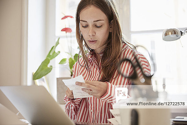 Young woman examining financial bill while using laptop at home