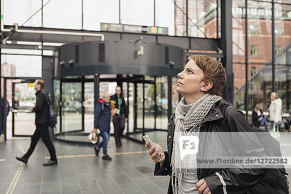 Thoughtful woman holding smart phone while looking up by building in city