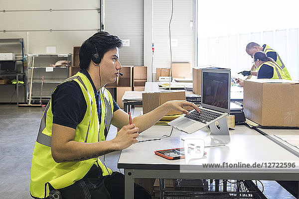 Confident young male customer service representative looking away while sitting with laptop at desk in warehouse