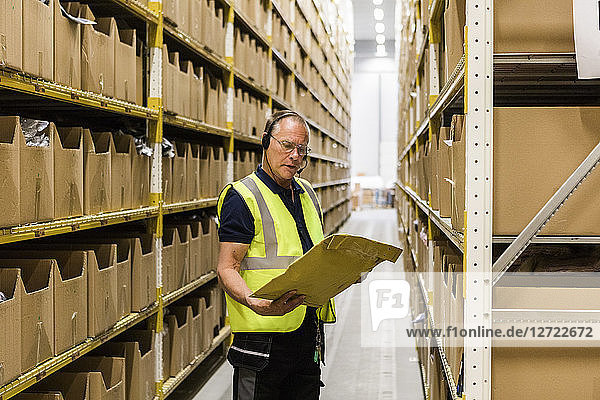 Senior male worker looking at package while talking through headset standing amidst racks at distribution warehouse