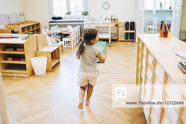 Full length rear view of girl running with book in classroom at kindergarten