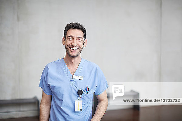 Portrait of smiling young male nurse in blue scrubs standing against wall at hospital