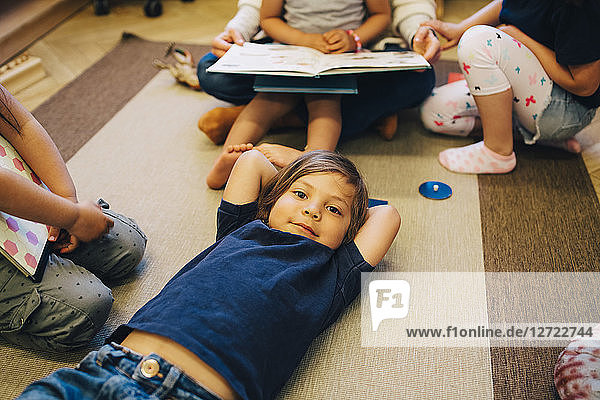 High angle portrait of boy lying on carpet amidst friends in classroom