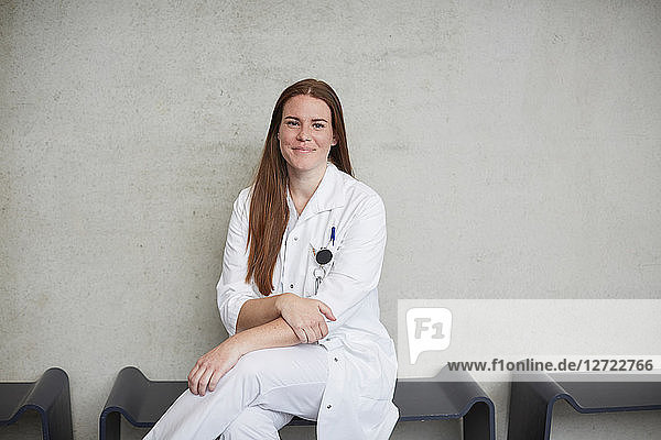 Portrait of smiling young female brunette doctor sitting against wall at hospital