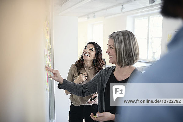 Mature female engineers smiling while reading adhesive notes stuck on glass in office