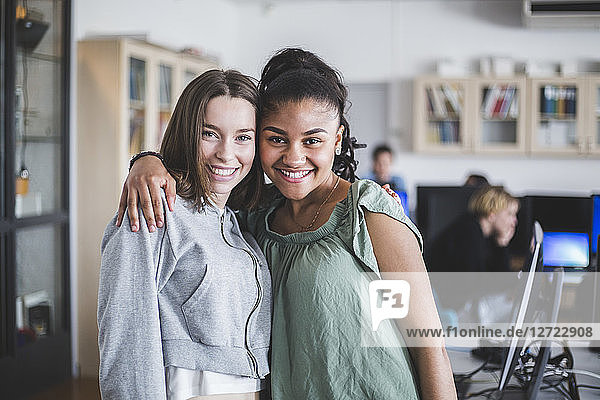 Portrait of smiling female teenage friends standing with arm around in computer lab at high school
