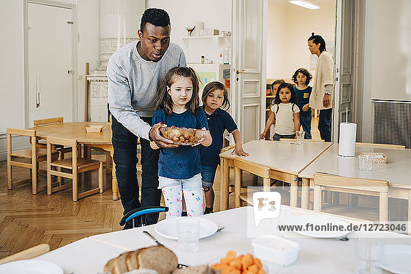 Male teacher assisting girl in carrying food bowl at child care classroom