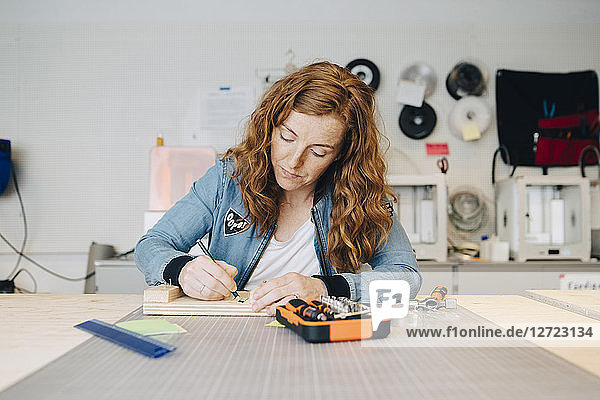 Confident redhead female engineer writing on wood at workbench in creative office