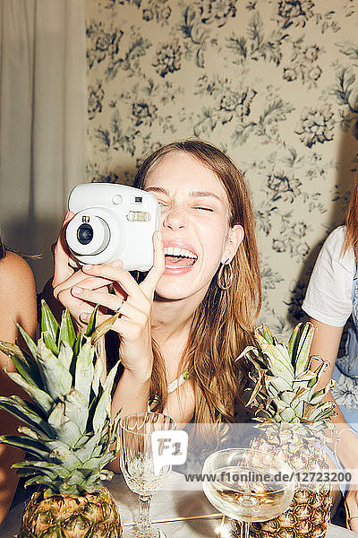 Cheerful young woman photographing while sitting amidst female friends at home during dinner party