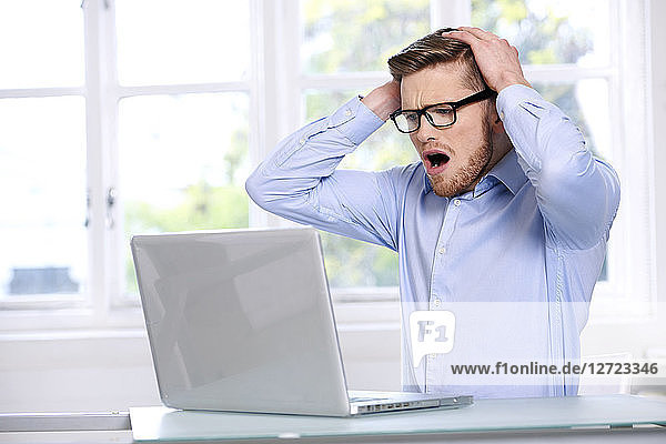 Mann in blauem Hemd  Brille  Bart  ernst  Fenster im Hintergrund unscharf  Mund geöffnet; Hände auf dem Kopf; sitzend  auf seinen Laptop blickend .