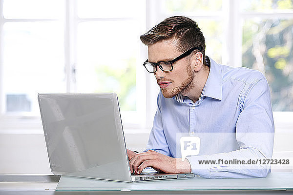 man in a blue shirt  glasses  beard  serious  window out of focus in the background  sitting  typing on a computer laptop